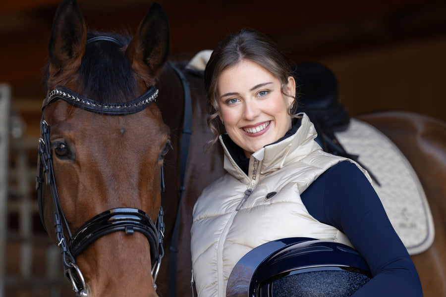 Cavalière en doudoune beige à l'écurie, casque sous le bras, à côté de son cheval