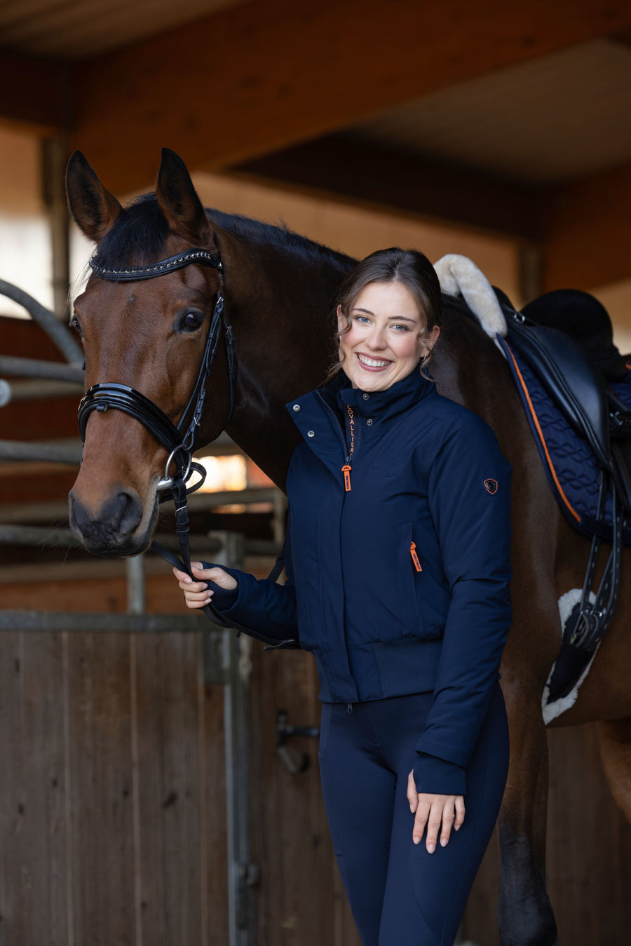 Cavalière en blouson d'hiver bleu marine à l'écurie, à côté de son cheval sellé