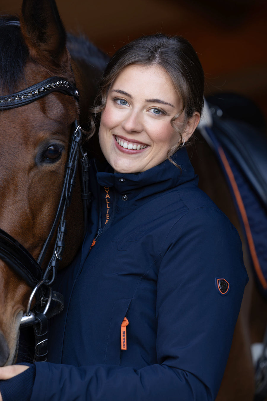 Cavalière souriante en blouson bleu marine, contre la tête de son cheval