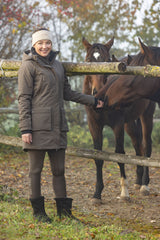 Cavalière en parka chocolat au lait appuyée à la clôture du pré, chevaux derrière